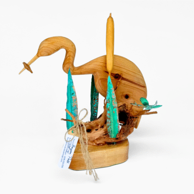Wooden heron sculpture on driftwood base. Shows long neck, beak, and light wood grain. Teal metal reeds and a small green insect evoke tranquil wetland art.