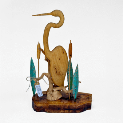Elegant hand-carved wooden heron sculpture with natural grain, posed on a polished base. Features stylized metal reeds with teal patina, evoking marsh artistry.